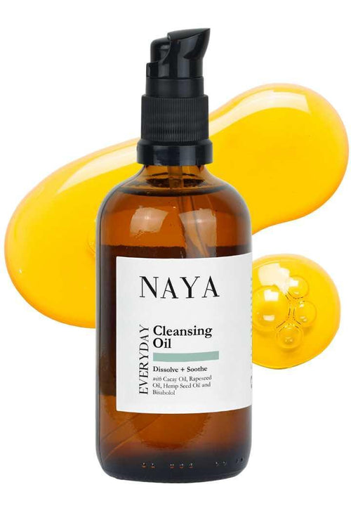 A bottle of NAYA Everyday Cleansing Oil with a pump dispenser, surrounded by a splash of yellow oil.