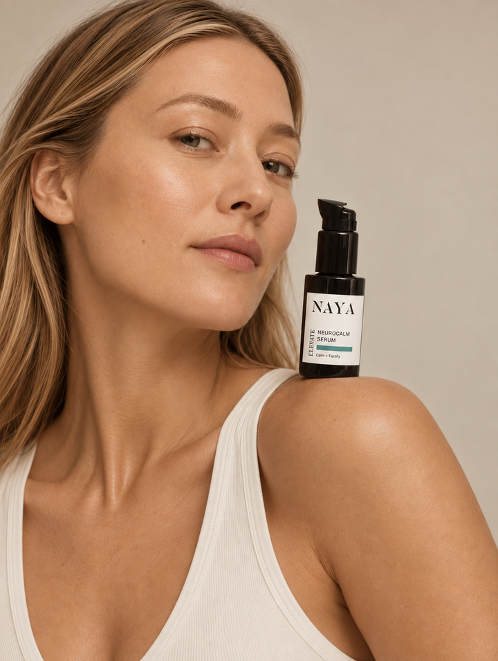 A close-up lifestyle shot of a woman with blonde hair wearing a white ribbed tank top, holding a black pump bottle of NAYA Skin Hydration Serum next to her shoulder against a neutral beige background.