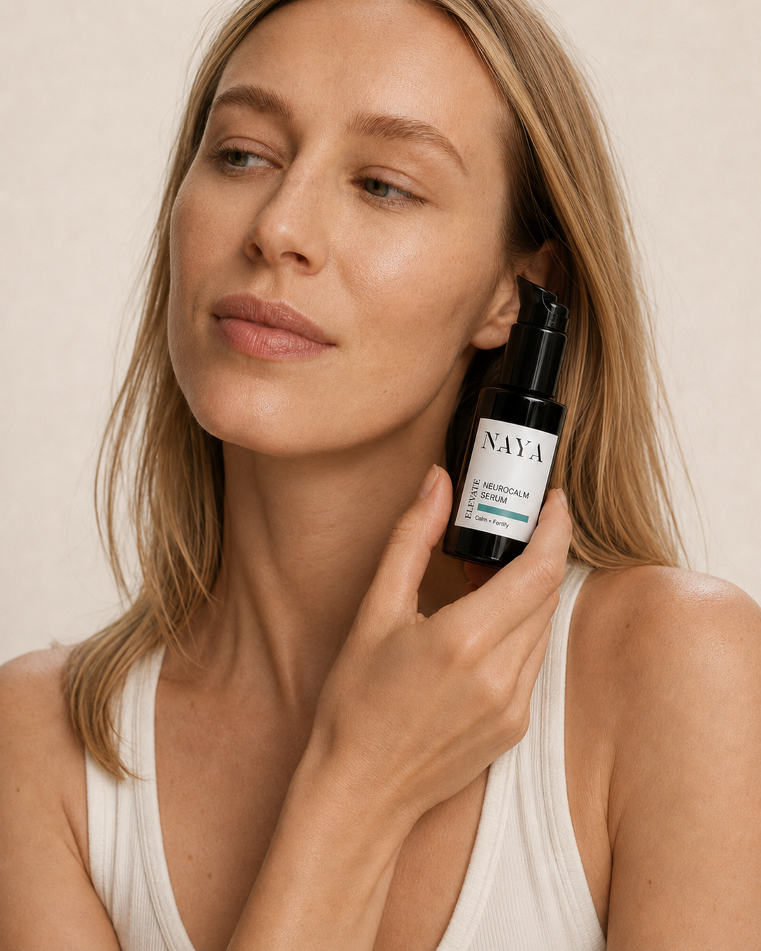 A woman with blonde hair holds a black pump bottle of NAVA Meristem Cream against her neck, wearing a white ribbed tank top against a warm beige background.